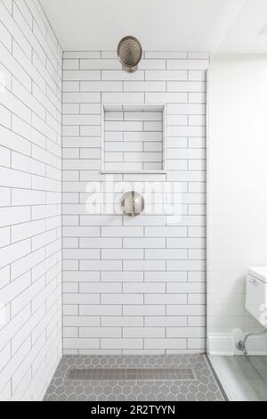 Eine Badezimmerdusche mit weißen Fliesenwänden aus der U-Bahn, ein eingebautes Nischenregal, ein Bronze-Duschkopf und ein grauer sechseckiger Fliesenboden. Stockfoto