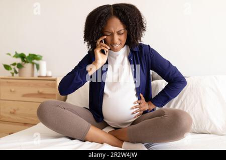 Verzweifelte, lockige, schwangere, schwarze Millenialfrau in Haushaltskleidung mit großen Bauchrufen per Telefon Stockfoto