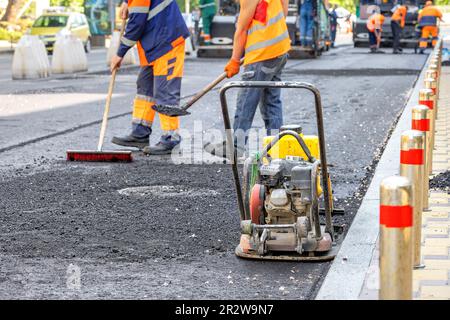 Eine alte verschlissene vibrierende Platte steht neben dem Bordstein vor dem Hintergrund einer Arbeiterbrigade von Straßenarbeitern, die frischen Asphalt verlegen. Speicherplatz kopieren. Stockfoto