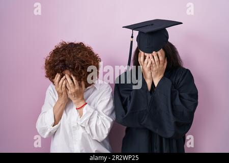 Hispanische Mutter und Tochter tragen Abschlussmütze und Zeremoniemäntel mit traurigem Ausdruck, der das Gesicht mit den Händen bedeckt, während sie weinen. Depressionskonzept. Stockfoto