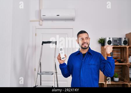Hispanischer Techniker, der mit Klimaanlage arbeitet, zeigt mit dem Daumen zur Seite und lächelt glücklich mit offenem Mund Stockfoto