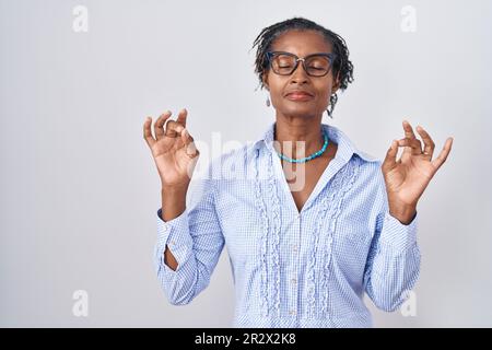 Afrikanische Frau mit Dreadlocks, die auf weißem Hintergrund steht und eine Brille trägt, entspannen sich und lächeln mit geschlossenen Augen und Meditationsgesten mit Fingern machen. Stockfoto