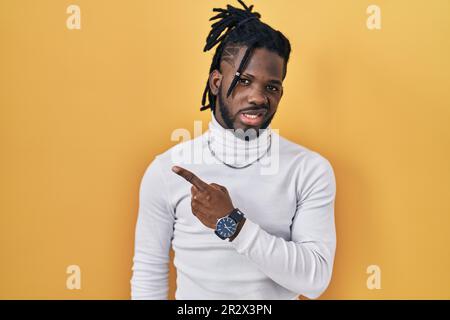 Ein afrikanischer Mann mit Dreadlocks, der einen Rollkragenpullover auf gelbem Hintergrund trägt und zur Seite zeigt, besorgt und nervös mit Zeigefinger, besorgt und surpri Stockfoto