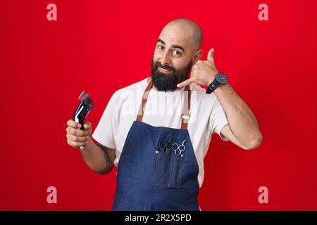 Junger lateinamerikanischer Mann mit Bart und Tätowierungen, der eine Barbier-Schürze trägt, mit einem lächelnden Rasiermesser, der Handygesten mit Hand und Fingern macht, als würde er über das Telefon reden Stockfoto