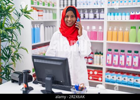 Eine junge, schöne Apothekerin, die mit einem Smartphone über einen Computer in der Apotheke spricht Stockfoto