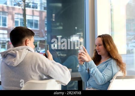 Das Leben von Teenagern am Telefon ein Junge und ein Mädchen sitzen mit ihrem Smartphone an einem Tisch am Fenster und verbringen Zeit im Internet in sozialen Netzwerken, wenn der wi-Fi-Hoodie grau weiß ist Stockfoto