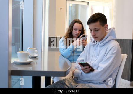 Das Leben von Teenagern am Telefon ein Junge und ein Mädchen sitzen mit ihrem Smartphone an einem Tisch am Fenster und verbringen Zeit im Internet in sozialen Netzwerken, wenn der wi-Fi-Hoodie grau weiß ist Stockfoto