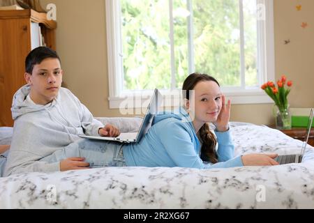 Das Leben von Teenagern auf einem Laptop ein Junge und ein Mädchen sitzen mit ihrem Smartphone an einem Tisch am Fenster und verbringen Zeit im Internet in sozialen Netzwerken, wenn der wi-Fi-Hoodie grau weiß ist Stockfoto