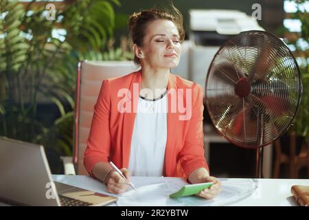 Nachhaltiger Arbeitsplatz. Lächelnde, moderne, 40 Jahre alte Buchhalterin in einem modernen grünen Büro in roter Jacke mit Taschenrechner, elektrischem Ventilator und Laptop. Stockfoto