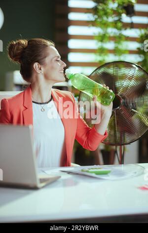 Nachhaltiger Arbeitsplatz. Glückliche, moderne 40-jährige Geschäftsfrau in einem modernen grünen Büro in roter Jacke mit einer Flasche Wasser und einem elektrischen Ventilator. Stockfoto