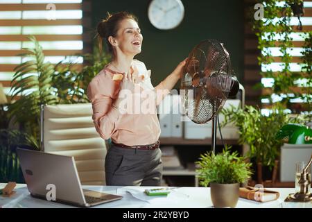 Nachhaltiger Arbeitsplatz. Glückliche, moderne, 40 Jahre alte Buchhalterin in einem modernen grünen Büro mit elektrischem Ventilator und Laptop. Stockfoto