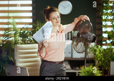 Nachhaltiger Arbeitsplatz. Glückliche, moderne 40 Jahre alte Mitarbeiterin in einem modernen grünen Büro mit Dokumenten und elektrischem Ventilator. Stockfoto