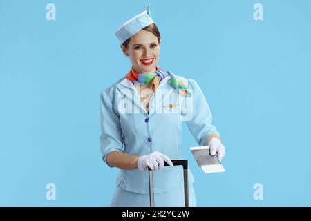 Lächelnde, stilvolle weibliche Stewardess auf blauem Hintergrund in blauer Uniform mit Rolltasche, Ticket und Pass. Stockfoto