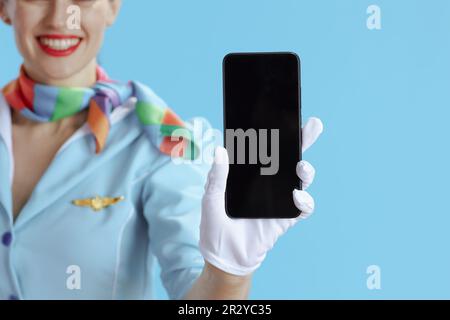 Nahaufnahme einer lächelnden weiblichen Stewardess vor blauem Hintergrund in blauer Uniform, die einen leeren Smartphone-Bildschirm zeigt. Stockfoto