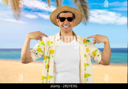 Ein lateinischer Mann im Urlaub, der am tropischen Strand eine Sonnenbrille mit geblümtem Hemd trägt und selbstbewusst mit einem Lächeln im Gesicht aussieht, sich mit stolzen Fingern zeigt Stockfoto