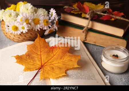 Buchen Sie mit Herbstblatt als Lesezeichen, Duftkerze und Kamillenblumen auf einem hellgrauen strukturierten Tisch, Nahaufnahme Stockfoto