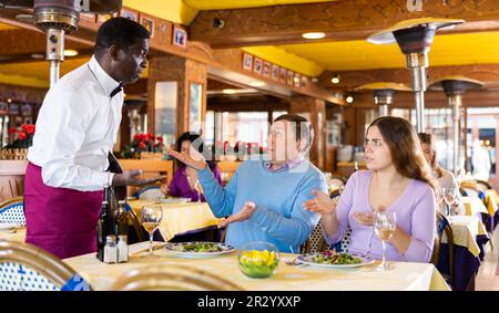 Unzufriedene Gäste, die mit dem afroamerikanischen Kellner im Restaurant in Konflikt geraten Stockfoto