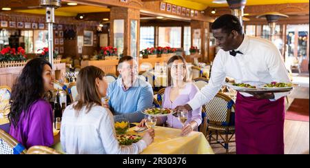 Ein afroamerikanischer Kellner serviert Speisen für eine fröhliche Gruppe von Erwachsenen im Restaurant Stockfoto