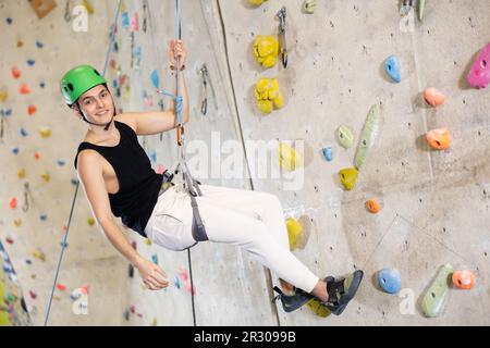 Gut ausgestatteter, positiver junger Mann, der am Seil runtergeht, nachdem er im Indoor Bouldering Adventure Park die Spitze erreicht hat Stockfoto