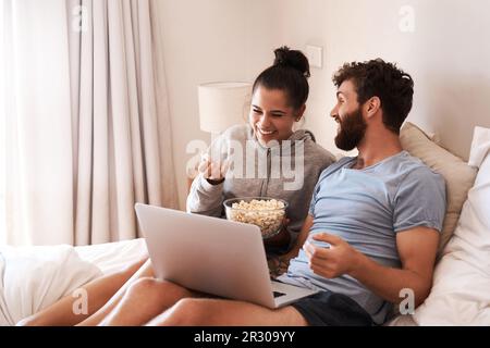 Laptop, Film und Popcorn mit lachenden Paaren im Bett zum Streamen, Entspannen oder Internet. Abonnement, glücklicher oder komischer Mann und Frau zu Hause Stockfoto