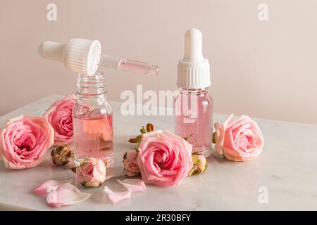 Eine offene Flasche Rosenöl und eine Pipette gefüllt mit Kosmetik auf einem Marmorpodium mit Blumen und Rosenblättern. Feuchtigkeitsspendend, weichmachend Stockfoto