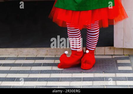 Rote, spitz zulaufende Schuhe aus Filz und gestreifte Strümpfe, Weihnachtself Mädchen Beine Fuß. Süße Füße in gestreiften roten weißen Strümpfen und lustigen Schuhen. Stockfoto