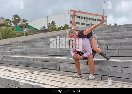 Ein Mann auf einer Treppe spielt, während er seinen Partner auf den Schultern trägt. Er hat einen weißen Bart und trägt dunkle Bermuda-Shorts. Sie trägt etwas Stockfoto