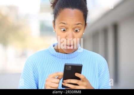 Vorderansicht Porträt einer überraschten schwarzen Frau, die auf der Straße ein Handy benutzt Stockfoto