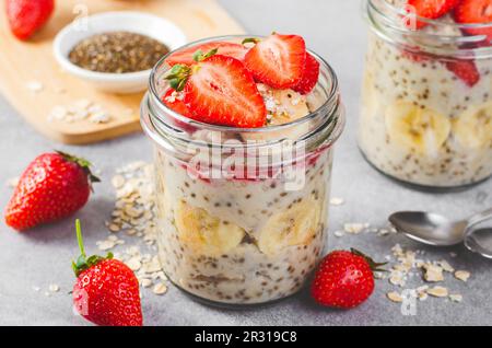 Haferflocken mit frischen Erdbeeren-, Bananen- und Chiasamen in Gläsern mit grauem Hintergrund, gesunder Snack oder Frühstück Stockfoto