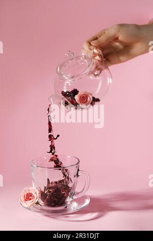 Frau gießt Hibiskus-Teeblätter aus der Teekanne in eine Tasse auf pinkfarbenem Hintergrund, Nahaufnahme Stockfoto