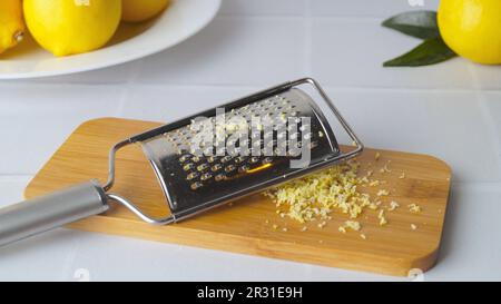 Holzbrett mit Reibe und frischer Zitronenschale auf einem weiß gefliesten Tisch Stockfoto