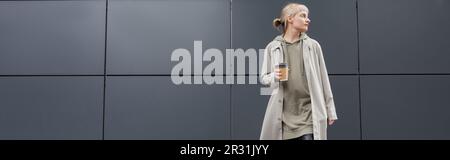 Eine stilvolle junge Frau mit Pony steht im Mantel und Kapuzenpulli und hält dabei einen Pappbecher mit Kaffee zum Mitnehmen in der Nähe eines grauen modernen Gebäudes an der Straße, draußen, Stockfoto