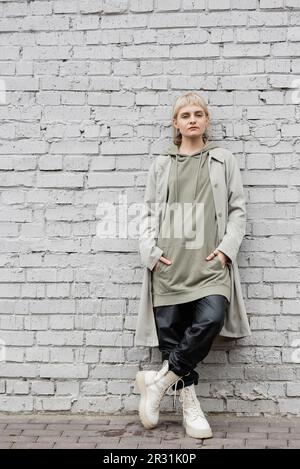 Junge Frau mit Make-up, blonden Haaren, Pony, in stilvollem Outfit, langer Kapuzenpullover, Mantel, schwarze Lederhosen und beige Stiefel stehen mit Händen in Taschen Stockfoto