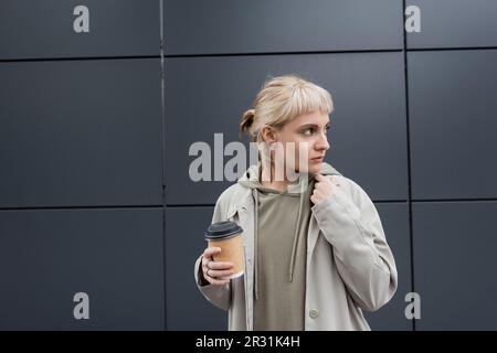 Modische junge Frau mit blonden Haaren und Pony steht im Mantel und Kapuzenpulli und hält Pappbecher mit Kaffee zum Mitnehmen in der Nähe eines grauen modernen Gebäudes Stockfoto