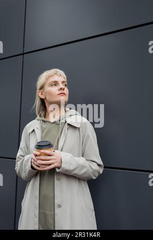 Junge Frau mit blonden Haaren und Knallen in stilvollem Outfit, Mantel und Kapuzenpulli, während sie einen Pappbecher mit Kaffee zum Mitnehmen in der Nähe einer grauen, modernen Bügel hält Stockfoto