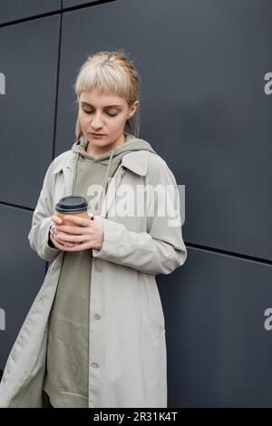 Junge Frau mit blonden Haaren und Pony stand im Mantel und Kapuzenpulli, während sie Pappbecher mit Kaffee zum Mitnehmen in der Nähe eines grauen modernen Gebäudes an der Straße hielt. Stockfoto
