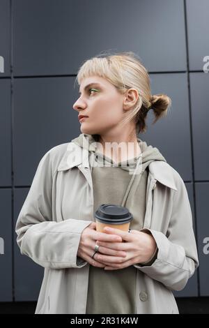 Stilvolle junge Frau mit blonden Haaren und Knallen, die im Mantel und Kapuzenpullover steht und dabei Pappbecher mit Kaffee zum Mitnehmen in der Nähe eines grauen modernen Gebäudes hält Stockfoto