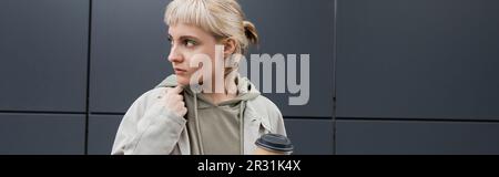 Stilvolle junge Frau mit blonden Haaren und Knallen, die im Mantel und Kapuzenpullover steht und dabei Pappbecher mit Kaffee zum Mitnehmen in der Nähe eines grauen modernen Gebäudes hält Stockfoto