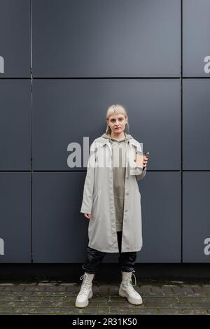 Stilvolle junge Frau mit blondem Haar und Knallgeräuschen steht im Mantel und Kapuzenpullover, hält einen Pappbecher mit Kaffee und schaut in die Kamera, fast in Grau modern Stockfoto