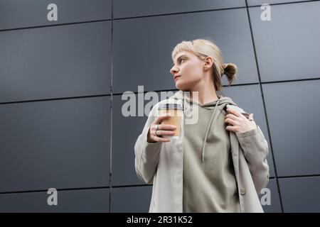 Stilvolle junge Frau mit blondem Haar und Pony, die wegsieht und in Mantel und Kapuze steht, während sie Pappbecher mit Kaffee zum Mitnehmen in der Nähe von Grey Mo hält Stockfoto