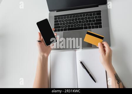 Ansicht von oben: Tätowierte Frau mit Kreditkarte und Smartphone, leerer Bildschirm neben Laptop und Notebook mit Stift auf weißem Tisch, moderner Arbeitsplatz, wo Stockfoto