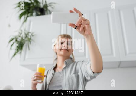 Blick aus dem niedrigen Winkel auf eine glückliche Frau mit Knallen und Ringen an den Fingern, die ein Glas Orangensaft halten und Selfie auf dem Smartphone machen und in verschwommenem Weiß stehen Stockfoto