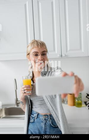Glückliche und tätowierte junge Frau mit Pony und Brille, die ein Glas Orangensaft hielt und Selfie auf einem verschwommenen Smartphone machte, während sie in Weiß stand Stockfoto