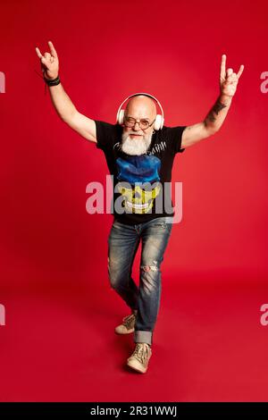 Rock 'n' Roll. Porträt in voller Länge emotionaler, reifer, bärtiger Mann mit Tattoos, der Musik in Kopfhörern vor rotem Studiohintergrund hört Stockfoto