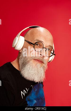 Nahaufnahme eines erwachsenen, bärtigen, glatzköpfigen Mannes mit Brille, der Musik über Kopfhörer hört und vor rotem Studiohintergrund in die Kamera schaut Stockfoto