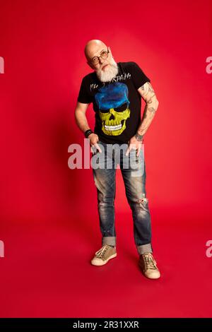 Porträt eines bärtigen, reifen, glatzköpfigen Mannes mit Brille und legerer, stylischer Kleidung mit Tattoos vor rotem Studiohintergrund Stockfoto