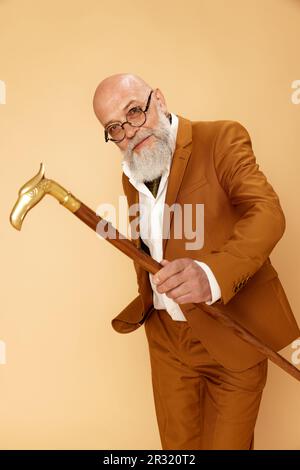 Porträt eines bärtigen, reifen, kahlen Mannes in elegantem, klassischem Anzug mit Spazierstock, der mit einem Lächeln vor Studiohintergrund posiert Stockfoto