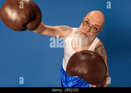 Porträt eines kahlköpfigen, bärtigen, reifen Mannes mit Tattoos in Sportbekleidung, der in alten Boxhandschuhen vor blauem Studiohintergrund posiert Stockfoto