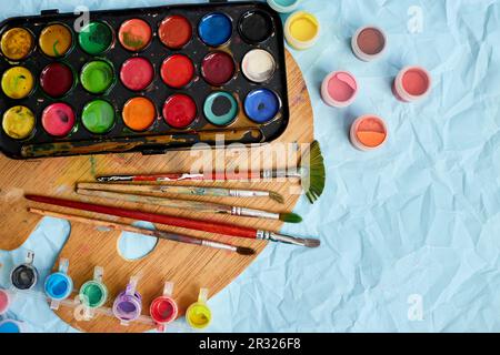 Flache Anordnung von bunten Farben, Malerpalette und Pinseln auf blauem Hintergrund, Kunst, Malen und Hobbykonzept, Draufsicht, Kopierbereich. Stockfoto