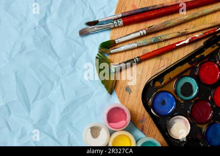 Flache Anordnung von bunten Farben, Malerpalette und Pinseln auf blauem Hintergrund, Kunst, Malen und Hobbykonzept, Draufsicht, Kopierbereich. Stockfoto